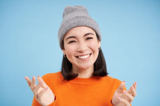 Close up portrait of happy smiling asian woman shows open hands, pointing, looking friendly, standing over blue background.