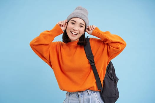 Stylish young asian woman in warm hat, walking with backpack, going somewhere with bag, standing over blue studio background.