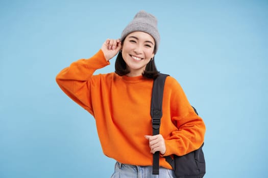 Cute smiling asian girl puts on warm hat to go outside, walks with backpack in orange sweater, blue background.
