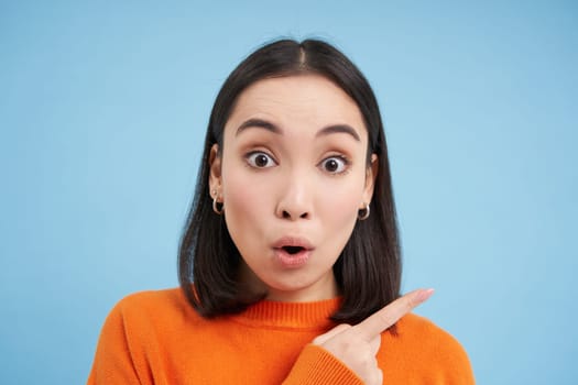 Close up of surprised asian girl, pointing finger right, showing advertisement with curious, intrigued face, standing over blue background.