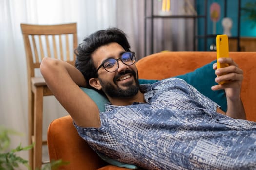 Indian man lying on couch uses mobile phone smiles at home living room apartment. Hindu young guy texting share messages content on smartphone social media applications online, watching relax movie