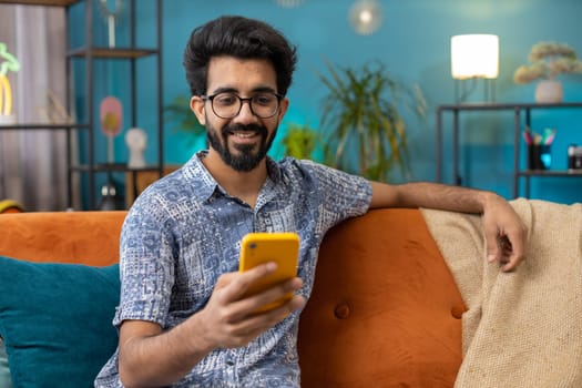 Indian man sitting on sofa uses mobile phone smiles at home living room apartment. Hindu young guy texting share messages content on smartphone social media applications online, watching relax movie