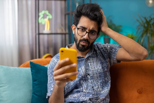 Sad displeased indian man use smartphone typing browsing, loses becoming surprised sudden lottery results, bad news, fortune loss, fail, deadline, virus. Young hindu guy at home living room on couch