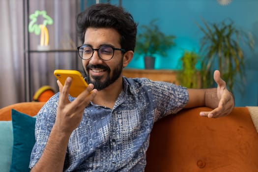 Happy excited indian man making online phone call conversation with friends. Portrait of hindu guy enjoying mobile loudspeaker talking chatting communication at home apartment living room on couch