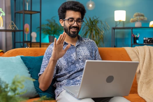 Indian man sitting on couch looking at camera, making video webcam conference call with friends or family, enjoying pleasant conversation. Young hindu guy laughing waving hello at home room