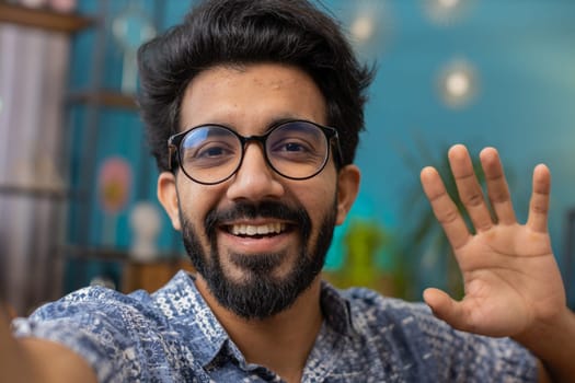 POV of indian man blogger taking selfie on smartphone, communicating video call online with social media subscribers followers, recording vlog stories. Young hindu guy at home apartment room on couch