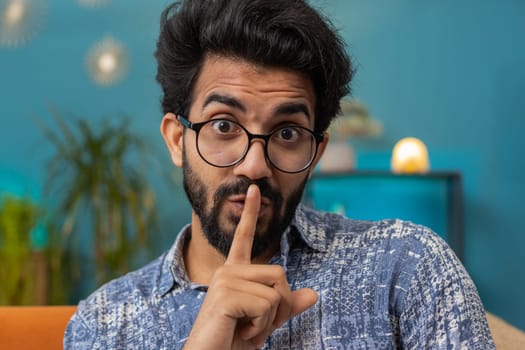 Shh be quiet, hush please. Indian man with presses index finger to lips makes silence hush gesture sign do not tells gossip secret. Young hindu guy at modern home living room sitting on couch. Gossip
