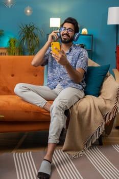 Happy overjoyed indian man in wireless headphones relaxing sitting on couch at home apartment choosing listening favorite energetic disco dancing music. People weekend daytime leisure activities