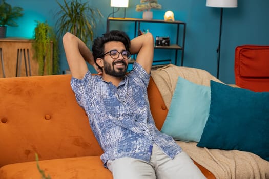 Tired indian man lying down on comfortable couch, taking a rest, daytime nap. Portrait of carefree hindu guy holding hands behind head relaxing, falling asleep at home apartment living room on sofa