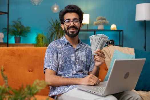 Planning budget. Rich happy indian man counting money cash use laptop computer calculate domestic bills at home room. Hindu guy satisfied of income earnings, saves money for planned vacation, gifts