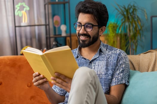 Young indian man relaxing interesting book, turning pages smiling enjoying literature, taking a rest on comfortable couch. Portrait of peaceful cheerful hindu guy at home apartment living room on sofa