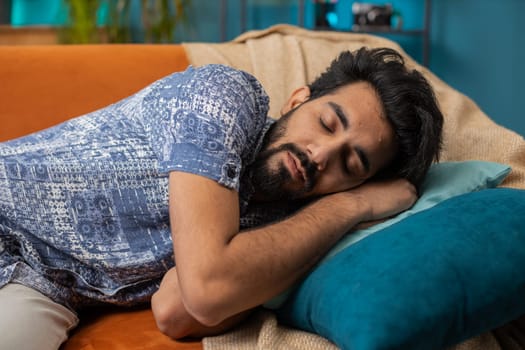 Tired indian man lying down in bed taking a rest. Carefree young hindu guy napping, falling asleep on comfortable sofa with pillows at home apartment room. Closed his eyes enjoy daytime nap alone