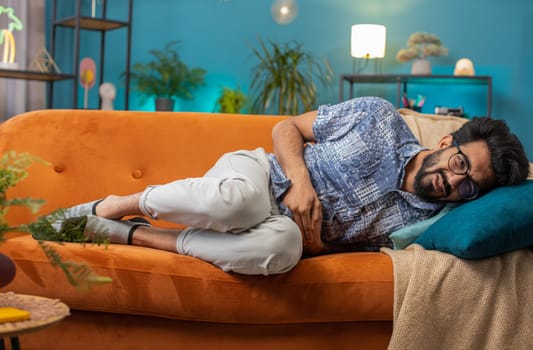 Young indian man lying on sofa feeling sudden strong abdominal stomach ache, gastritis problem. Hindu guy having symptom poisoning diarrhea indigestion peptic ulcer pancreatitis at home apartment room