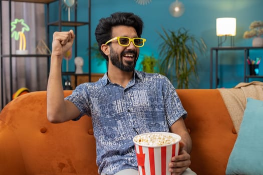 Excited indian man in 3D glasses sits on couch eating popcorn snacks and watching interesting TV serial sport game, film online social media movie content at home apartment. Guy enjoying entertainment