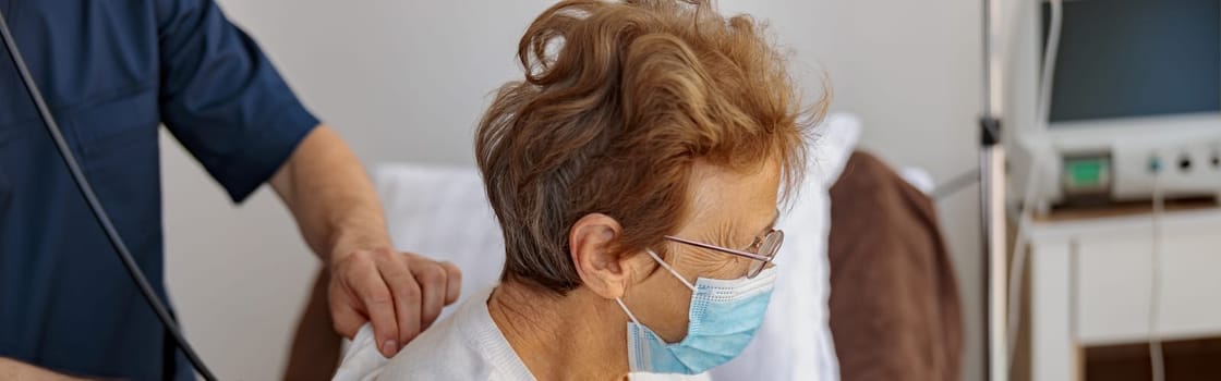 Doctor listening to old woman's patient breathing, using stethoscope in hospital ward