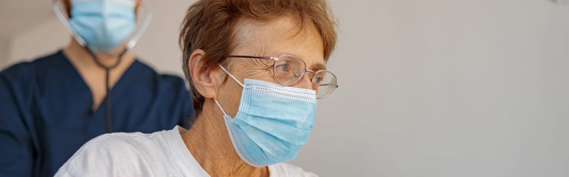 Doctor in face mask listening to old woman's patient breathing, using stethoscope in hospital ward