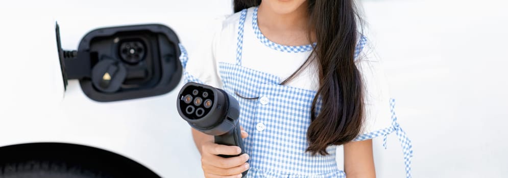 A playful girl holding and pointing an EV plug, a home charging station providing a sustainable power source for electric vehicles. Concept of progressive new generation with ecological awareness.