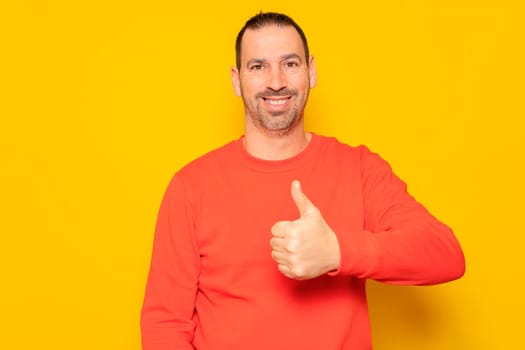 Bearded Hispanic man in his 40s wearing a red jumper proudly raising his thumb up while looking and smiling at the camera, isolated on yellow studio background