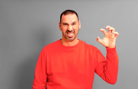 Bearded hispanic man in his 40s wearing a sweater looking extremely angry showing his claw hand while having an aggressive and defiant expression, isolated over gray background.
