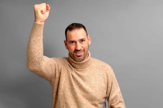 Bearded Hispanic man in his 40s wearing a beige turtleneck raising his fist energetically celebrating a great victory, isolated over gray background.