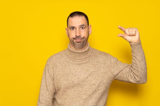 Bearded Hispanic man in his 40s wearing a turtleneck sweater gesturing small or little amount with a disgusted grimace on his face, isolated on yellow studio background.