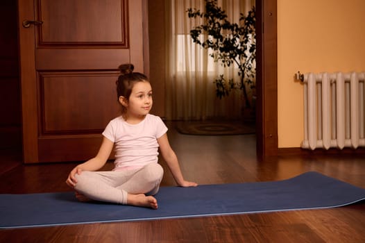 Adorable lovely Caucasian 5 years old little child girl in active wear, smiling, looking away, sitting on yoga mat at cozy home interior. Happy children. Childhood. Sport, lifestyle and hobby concept
