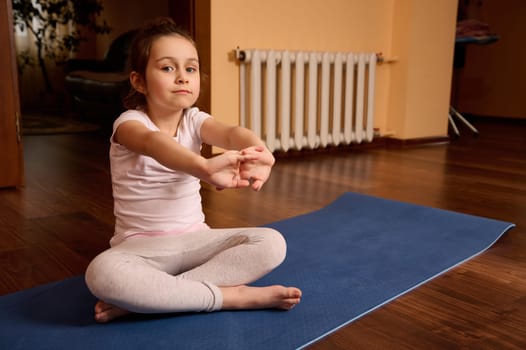 Caucasian lovely kid girl 5 years old, sitting barefoot on yoga mat, stretching arms, performing morning workout at home interior, looking confidently at camera. Sport. Fitness. Active child