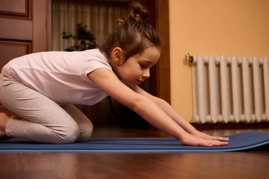 Close-up little girl in sportswear, stretching her body in Balasana pose, sitting in child pose while practicing yoga on a blue fitness mat in a cozy wooden home interior. Healthy habits. Childhood