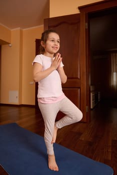 Full length portrait of a beautiful smiling girl child practicing yoga, standing in tree pose on blue fitness mat, working out, wearing sportswear, white t-shirt, and light gray leggins. Meditation.