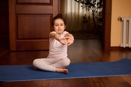 Caucasian lovely little child girl sitting on fitness mat, stretching her arms while exercising, practicing morning yoga at home, looking at camera. Body care. Mindfulness.Active and healthy lifestyle