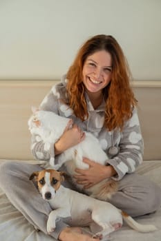 Caucasian woman holding a white fluffy cat and Jack Russell Terrier dog while sitting on the bed. The red-haired girl hugs with pets