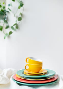 Empty yellow cups and brightly colored plates on a white table covered with a linen tablecloth, a flower pot on the wall in the background. Vertical photo