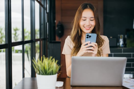 Happy lifestyle female smiling using smartphone in coffee shop, young business woman working with laptop computer she holding smart mobile phone at cefa for texting messages