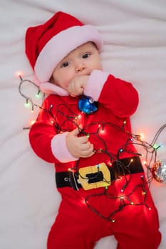 A baby boy dressed in a santa suit lies on the bed and plays with a Christmas garland