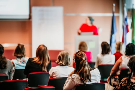 Business and entrepreneurship symposium. Female speaker giving a talk at business meeting. Audience in conference hall. Rear view of unrecognized participant in audience.