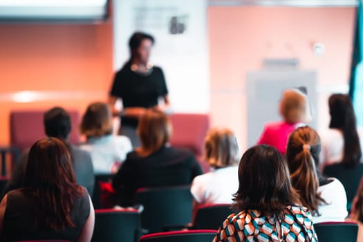 Business and entrepreneurship symposium. Female speaker giving a talk at business meeting. Audience in conference hall. Rear view of unrecognized participant in audience.