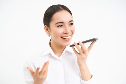 Professional businesswoman, asian lady smiles, speaks on speakerphone, records voice message on smartphone, white background.