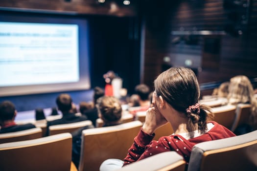 Business and entrepreneurship symposium. Female speaker giving a talk at business meeting. Audience in conference hall. Rear view of unrecognized participant in audience.