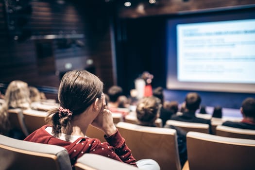 Business and entrepreneurship symposium. Female speaker giving a talk at business meeting. Audience in conference hall. Rear view of unrecognized participant in audience.