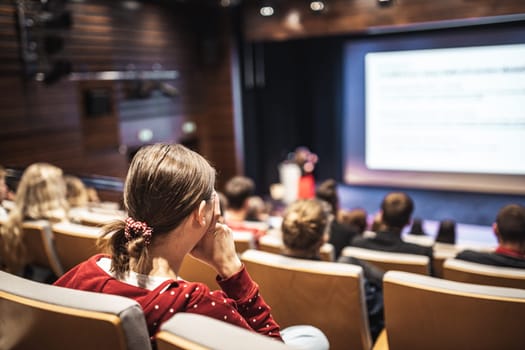 Business and entrepreneurship symposium. Female speaker giving a talk at business meeting. Audience in conference hall. Rear view of unrecognized participant in audience.