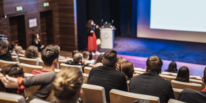 Business and entrepreneurship symposium. Female speaker giving a talk at business meeting. Audience in conference hall. Rear view of unrecognized participant in audience.