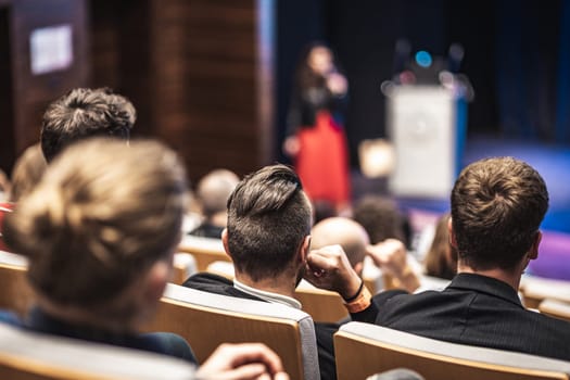 Business and entrepreneurship symposium. Female speaker giving a talk at business meeting. Audience in conference hall. Rear view of unrecognized participant in audience.