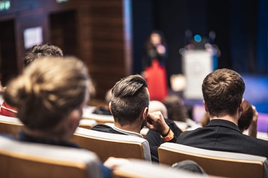 Business and entrepreneurship symposium. Female speaker giving a talk at business meeting. Audience in conference hall. Rear view of unrecognized participant in audience.