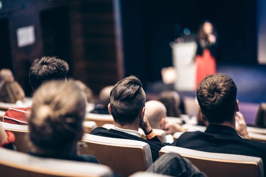 Business and entrepreneurship symposium. Female speaker giving a talk at business meeting. Audience in conference hall. Rear view of unrecognized participant in audience.