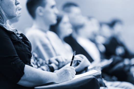 Female hands holding pen and notebook, making notes at conference lecture. Event participants in conference hall. Blue toned greyscale image