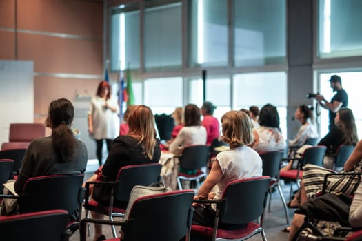 Business and entrepreneurship symposium. Female speaker giving a talk at business meeting. Audience in conference hall. Rear view of unrecognized participant in audience.