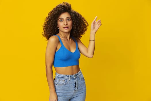 Woman with curly afro hair in a blue t-shirt on. yellow background signs with her hands, look into the camera, smile with teeth and happiness, copy space. High quality photo