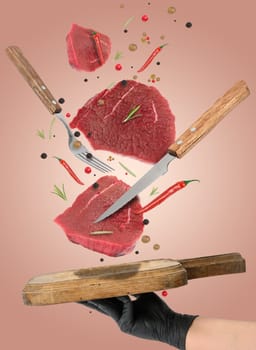 Raw pieces of beef, rosemary, spices and red chili peppers fall on a wooden cutting board