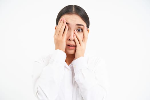 Japanese woman shuts face with hands, peeks through fingers with scared, terrified face, stands over white background.