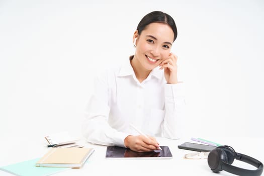 Portrait of asian woman in headphones, working on digital tablet, signing digital contracts, sitting in office and smiling, white background.